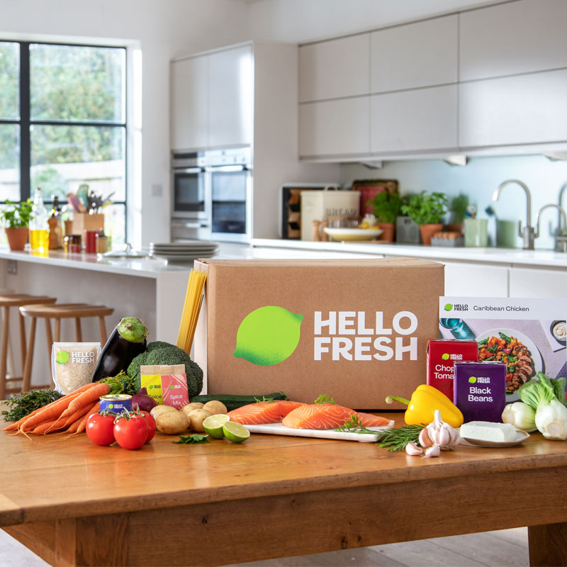 A HelloFresh box on a table surrounded by vegetables, fish, rice, pasta and a recipe card.