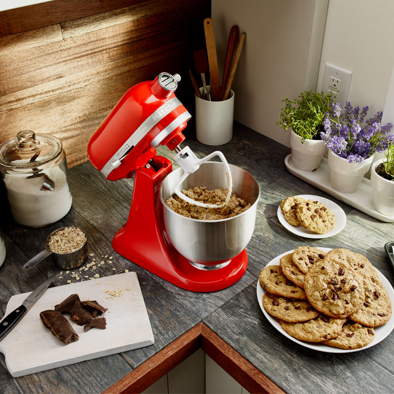 A red KitchenAid stand mixer on a kitchen counter surrounded by baking ingredients and plates of cookies.