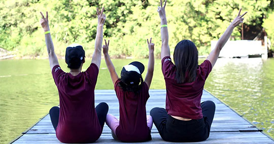Three people sit cross legged on a dock with their backs to the viewer making peace signs with their hands. 