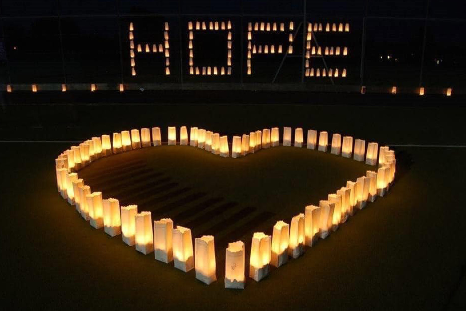 Glowing luminaries on the ground in the shape of a heart. Luminaries in the background spell out the word hope.